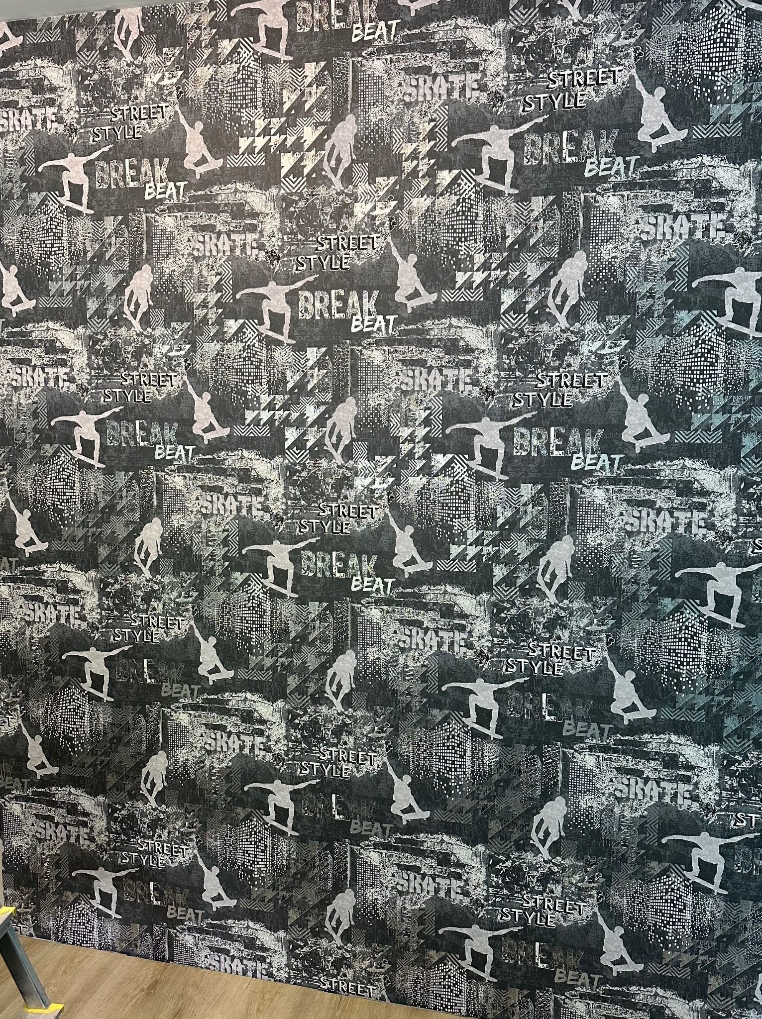 Ściana pokryta tapetą z motywem skateboardingu w stylu graffiti, z widocznym fragmentem podłogi i drabiny malarskiej w lewym dolnym rogu.