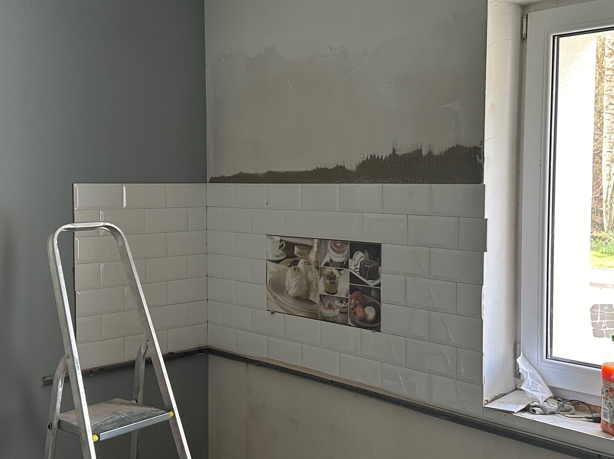 Ściana łazienki w trakcie remontu: białe, prostokątne płytki ułożone do połowy wysokości, z dekoracyjnym panelem z motywem deserów, widoczna drabina malarska i okno z resztkami materiałów budowlanych.