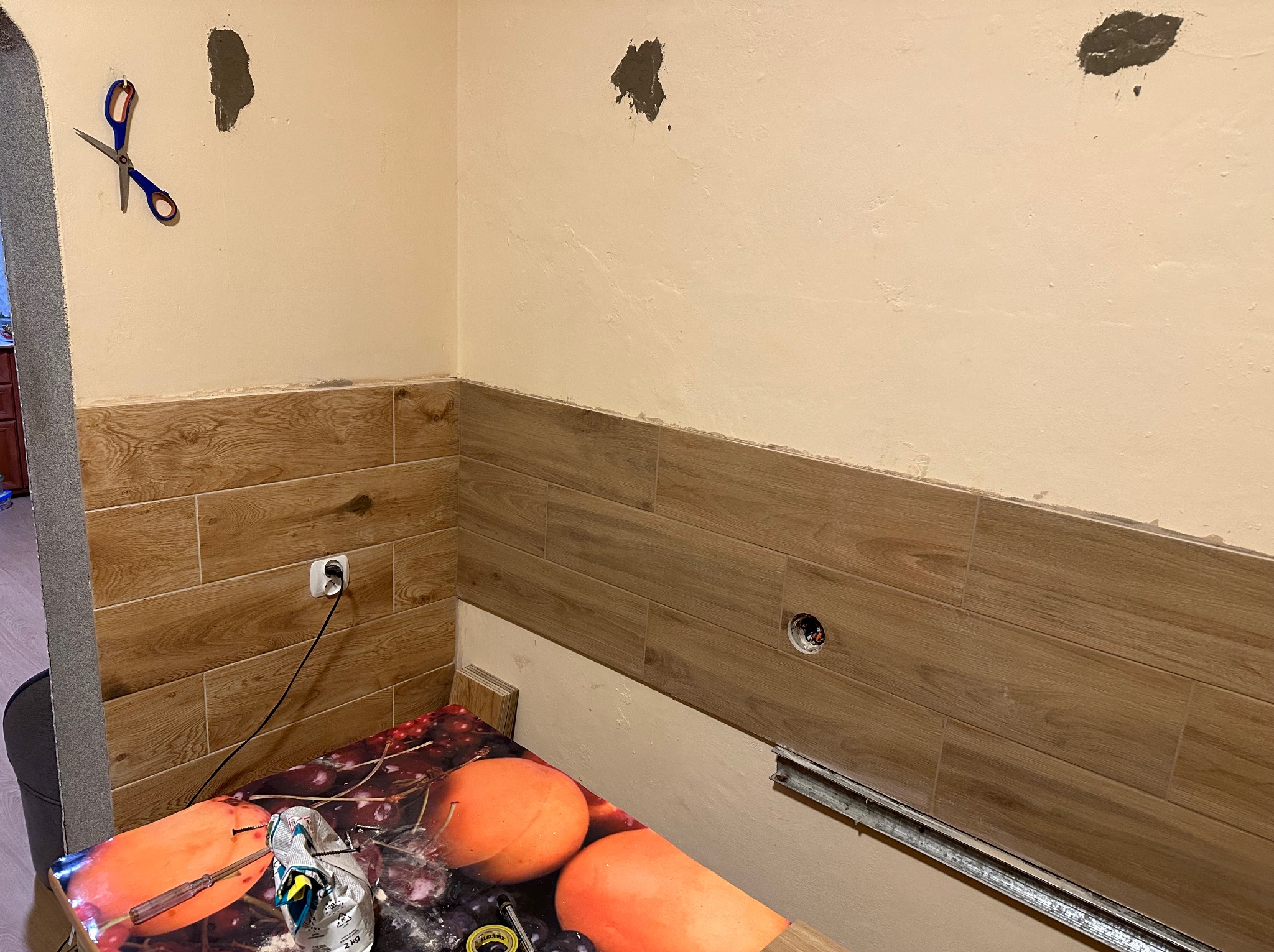 Ściana w trakcie układania płytek imitujących drewno, widoczne gniazdko elektryczne, puszka pod kontakt, szpachlowane ubytki w ścianie, nożyczki wiszące na ścianie, stół z narzędziami i zaprawą.