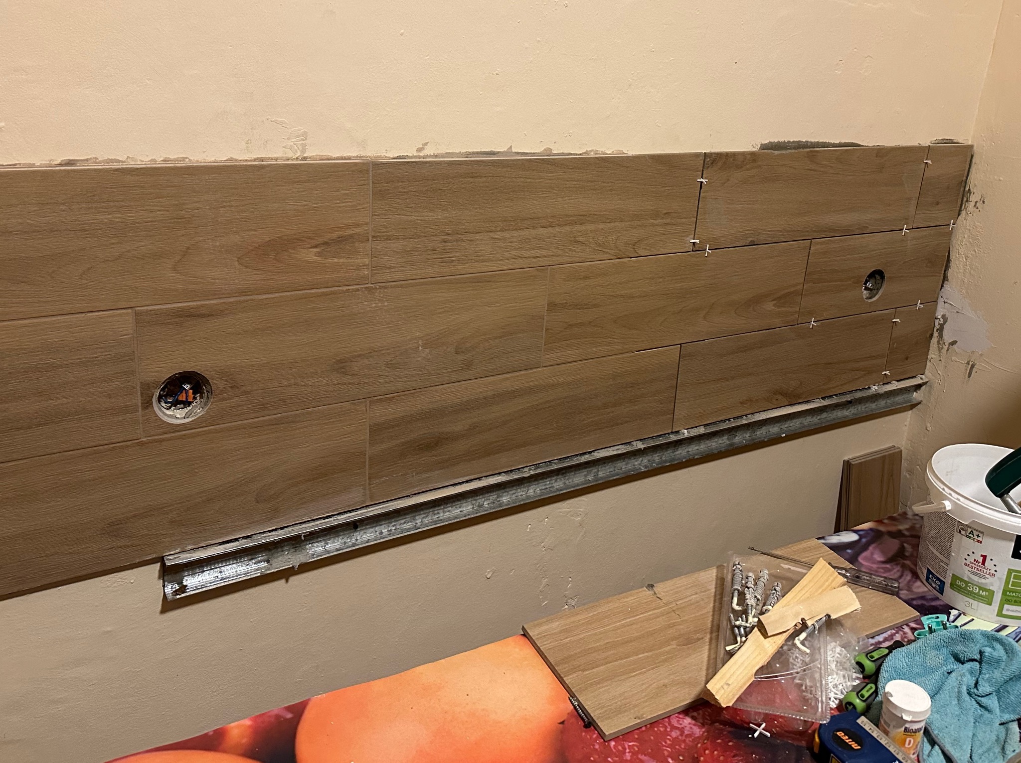 Montaż płytek imitujących drewno na ścianie z widocznymi otworami na gniazdka elektryczne, listwa montażowa i narzędzia glazurnicze na pierwszym planie.