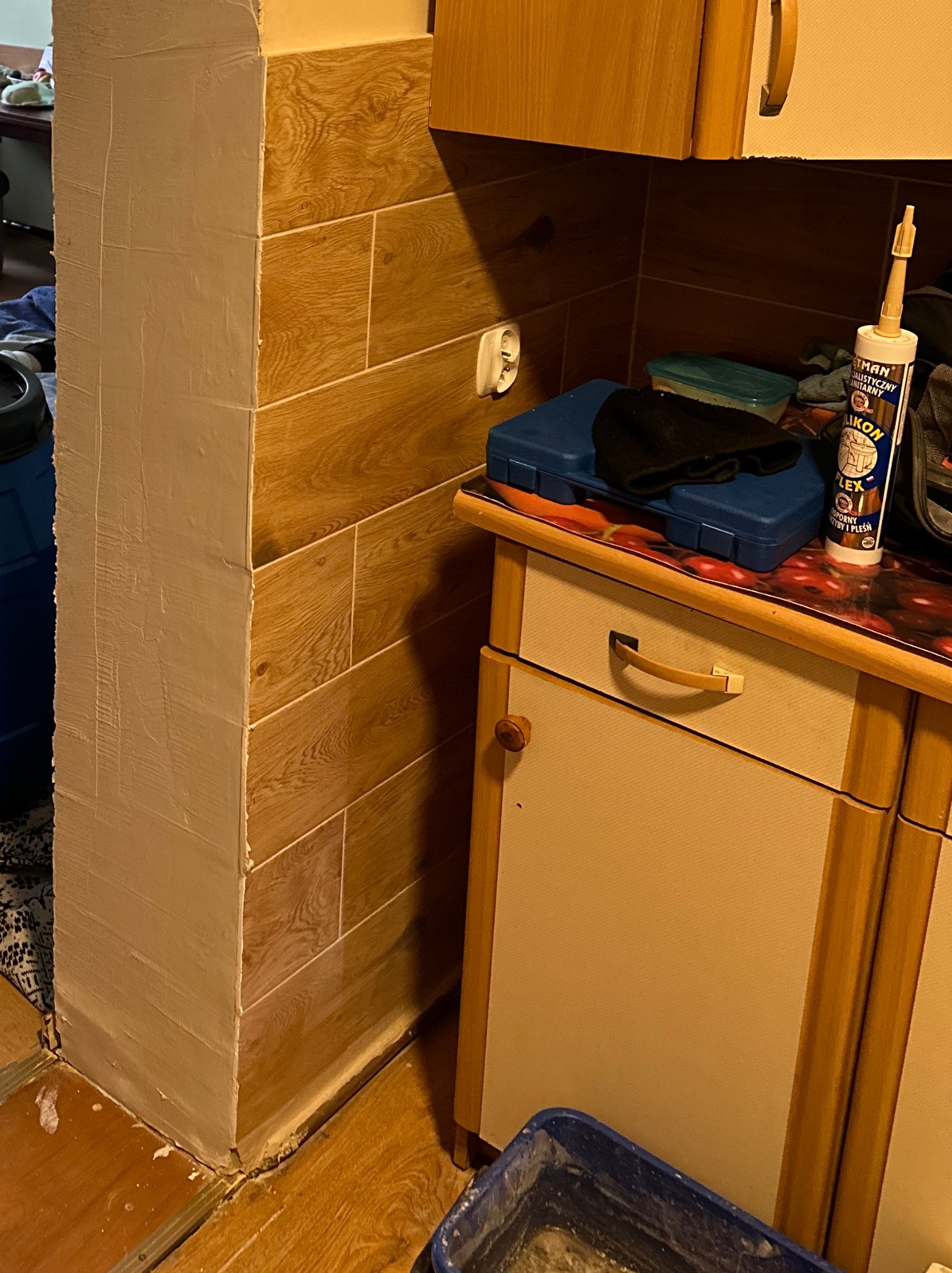 Obłożony płytkami imitującymi drewno filar, częściowo pokryty gładzią, widoczny silikon i narzędzia na blacie szafki.