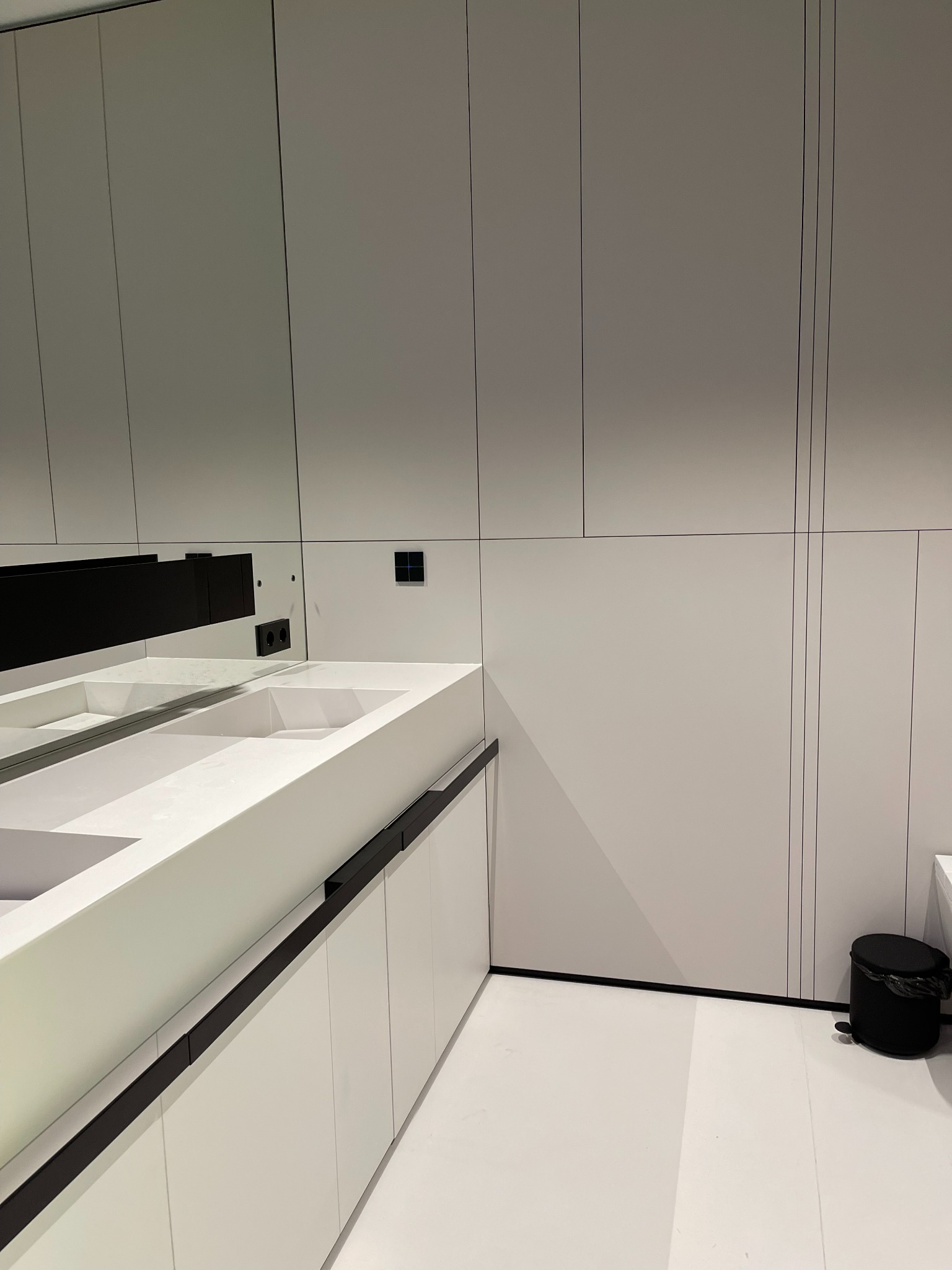 Nowoczesna łazienka z podwójną, białą umywalką nablatową o geometrycznych kształtach, zamontowaną na szafkach z czarnymi uchwytami. Ściana zabudowana białymi szafkami sięgającymi sufitu...