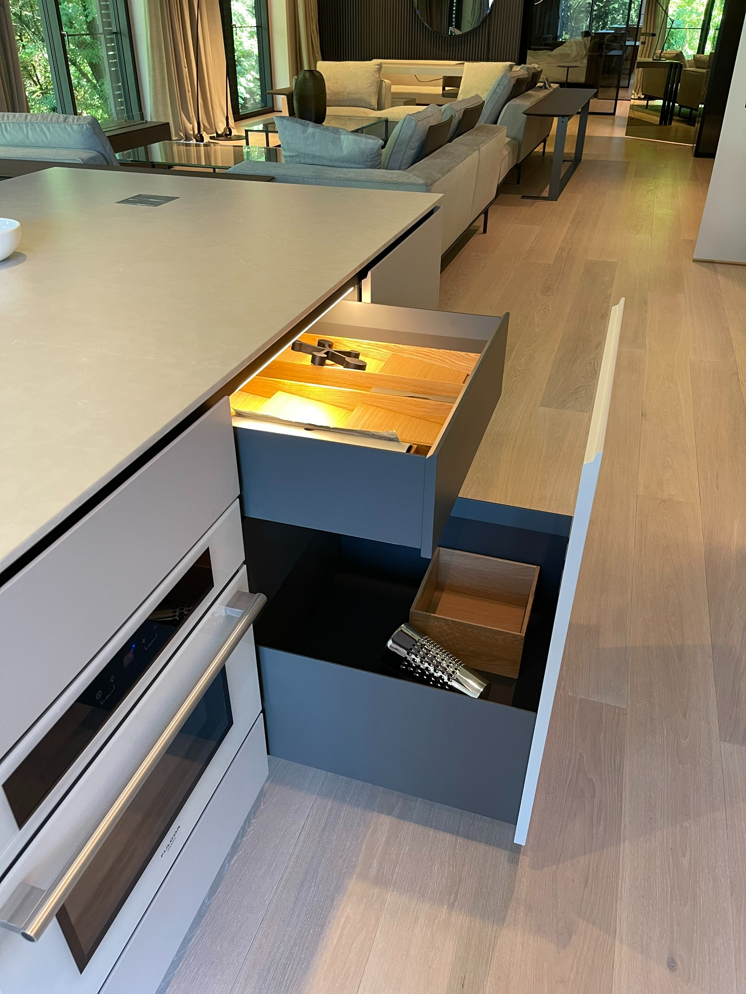 Szary blat kuchenny z wysuwanymi szufladami, jedna z drewnianym organizerem na sztućce, druga z ozdobnym wazonem, w tle salon z szarą sofą.