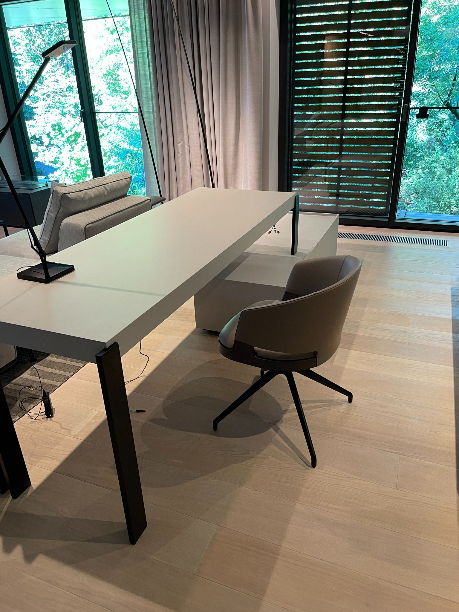 Nowoczesne biuro z długim, białym biurkiem z czarnymi nogami, obrotowym krzesłem w kolorze taupe i widokiem na zieleń przez okno z żaluzjami.