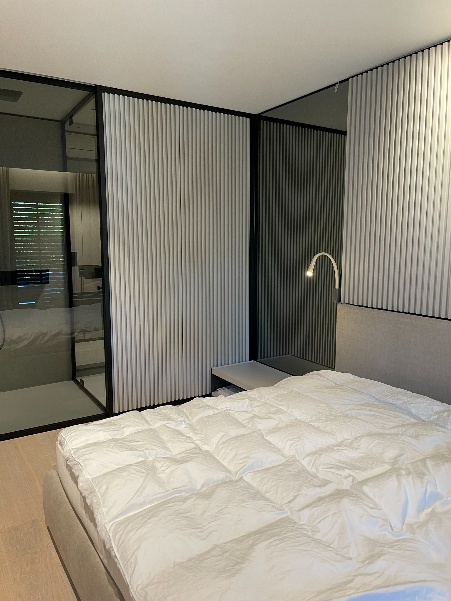 Nowoczesna sypialnia z łóżkiem z szarą ramą i białą pościelą, na tle ściany z pionowych paneli dekoracyjnych i przesuwanych drzwi do garderoby z czarnymi ramami.