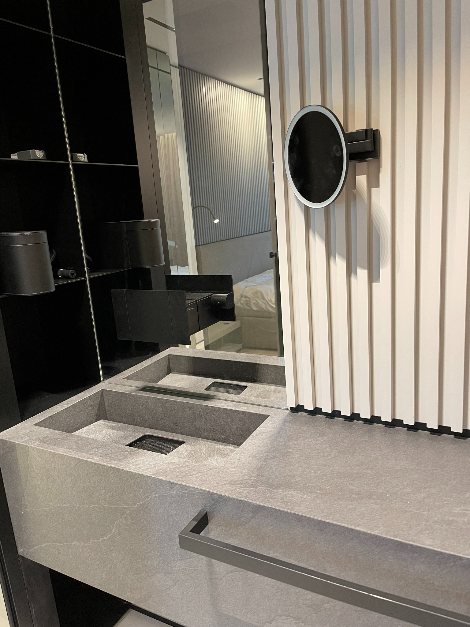 Nowoczesna łazienka z umywalką z szarego kamienia, okrągłym podświetlanym lustrem i czarnymi półkami w tle.