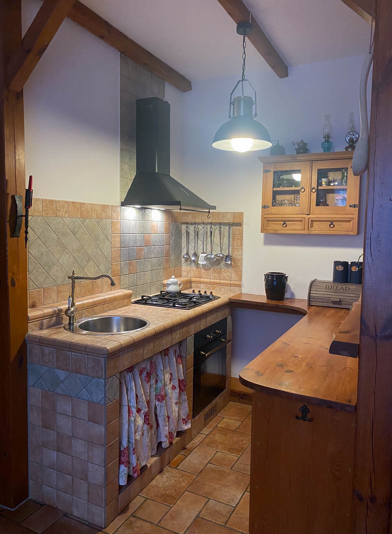 Rustykalna kuchnia z ceramiczną zabudową zlewu i blatu, okapem nad płytą gazową, drewnianymi belkami stropowymi i wiszącą lampą w stylu industrialnym.