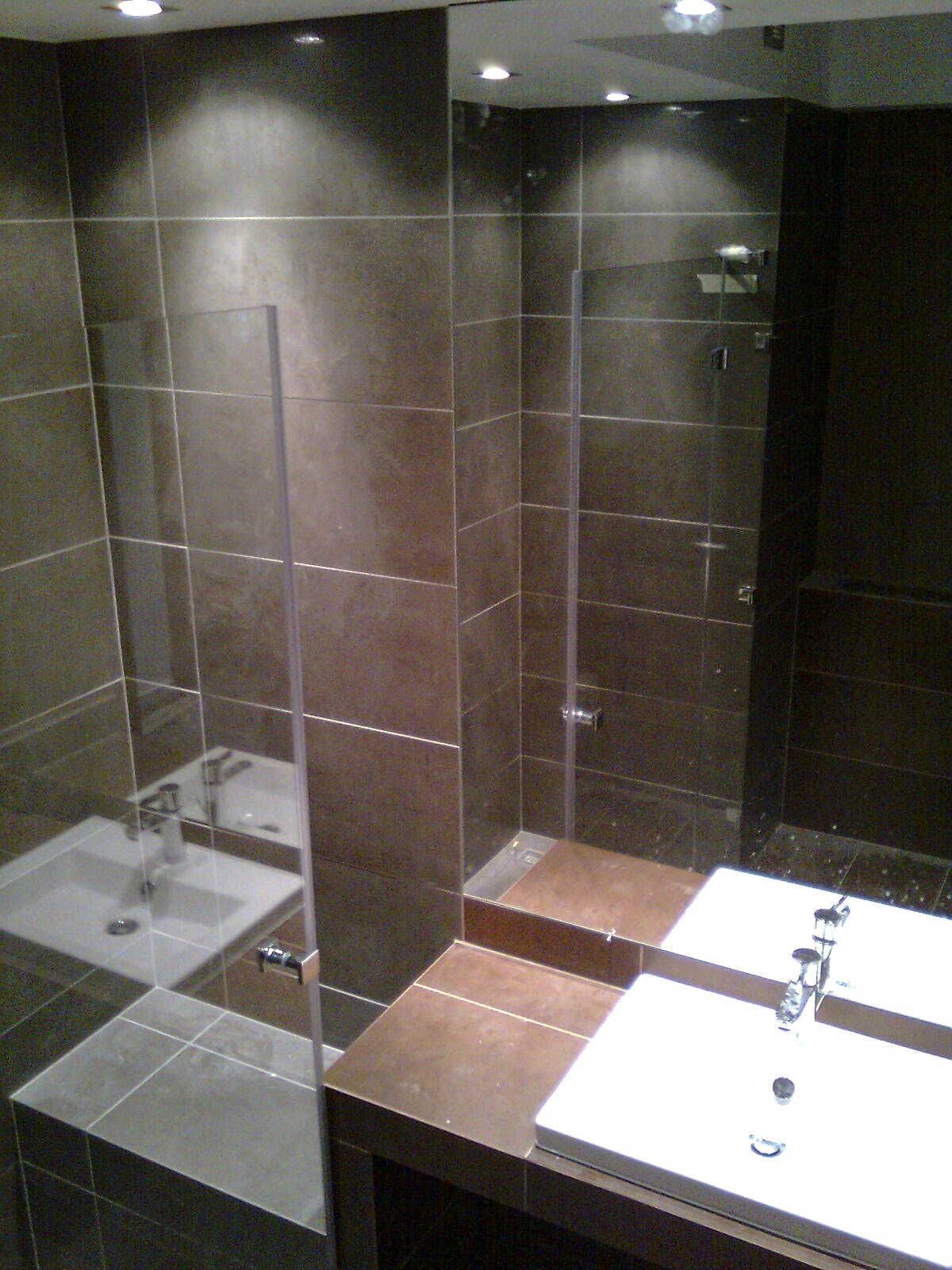 Nowoczesna łazienka z ciemnymi, kwadratowymi płytkami, kabiną prysznicową ze szklaną ścianką oraz podwójną, białą umywalką z chromowanymi bateriami, odbijająca się w dużym lustrze.