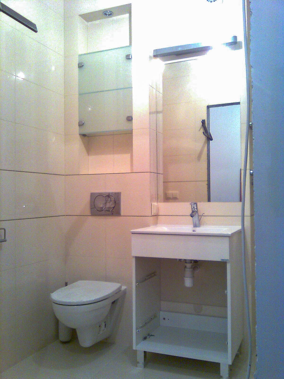 Jasna łazienka z wiszącą toaletą, umywalką z szafką bez frontów szuflad i lustrem, widoczne elementy instalacji hydraulicznej pod umywalką.