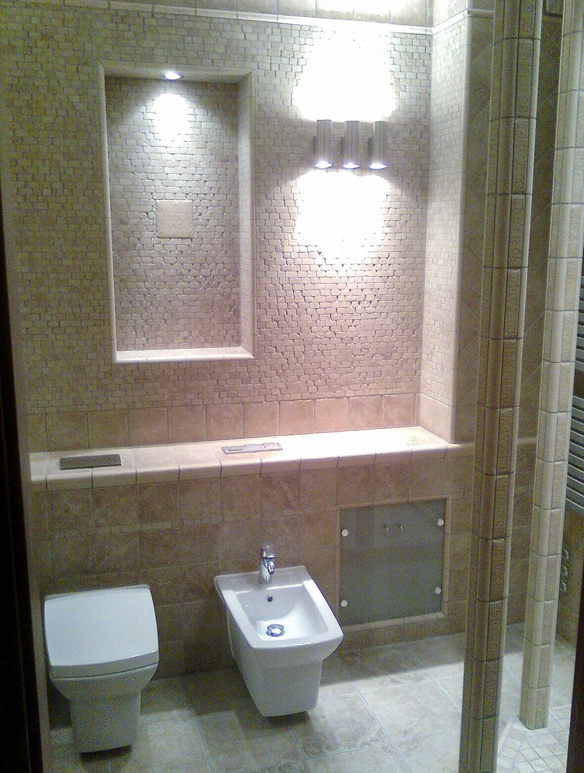 Jasna łazienka z mozaikową ścianą, wnęką z oświetleniem punktowym i nowoczesną armaturą: toaleta i bidet.