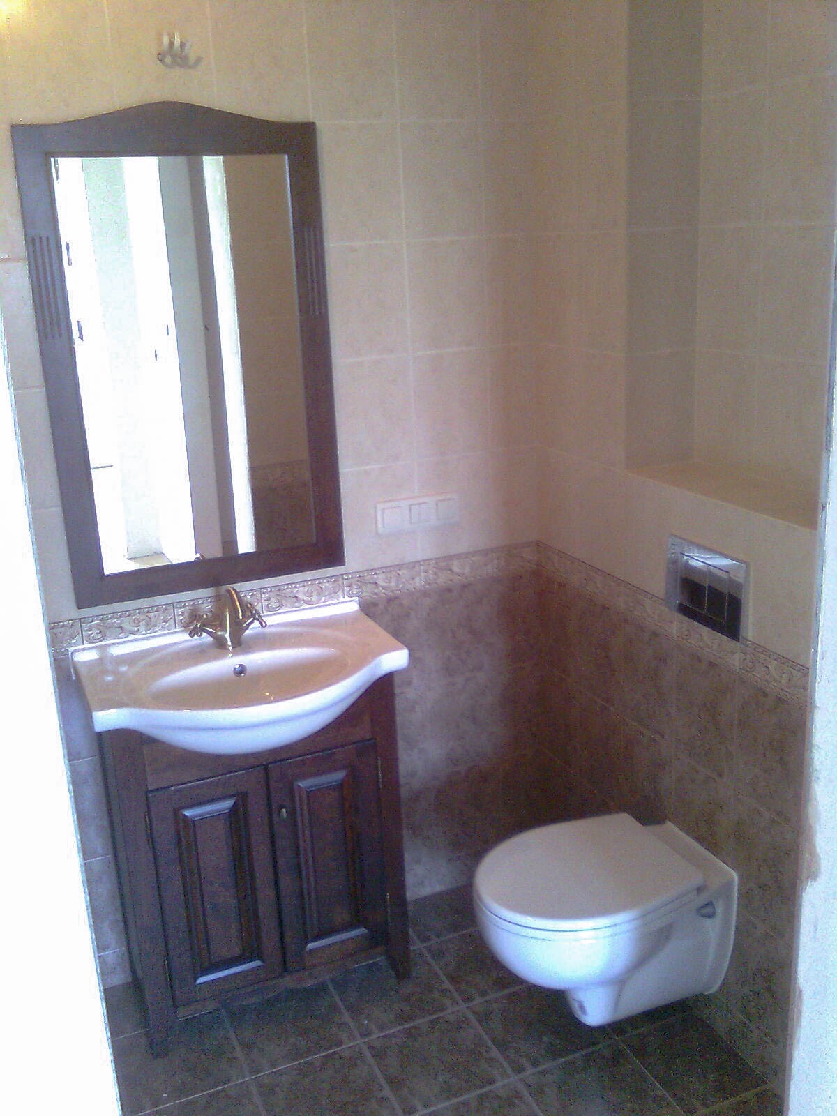 Wykończona łazienka z brązowymi płytkami, umywalką na szafce z ciemnego drewna, złotą baterią i wiszącą toaletą