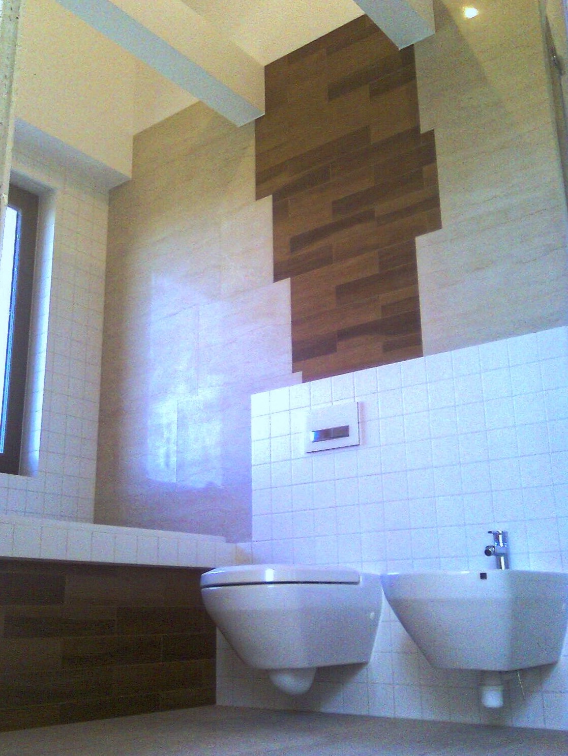 Nowoczesna łazienka z podwieszaną toaletą i bidetem na białej, kafelkowanej ścianie, uzupełniona o dekoracyjny panel z płytek imitujących drewno i jasne, ceramiczne wykończenie ścian.