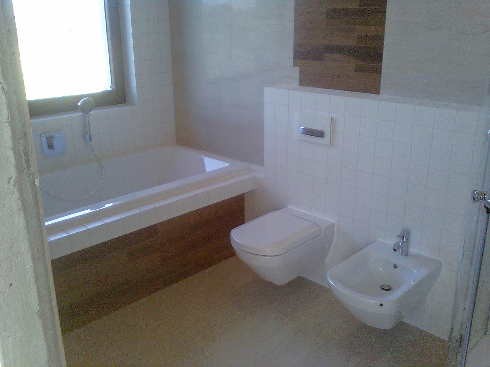 Nowoczesna łazienka z białą wanną obudowaną drewnopodobnymi płytkami, podwieszaną toaletą i bidetem na tle białych, kwadratowych płytek. Widoczny fragment kabiny prysznicowej po prawej.