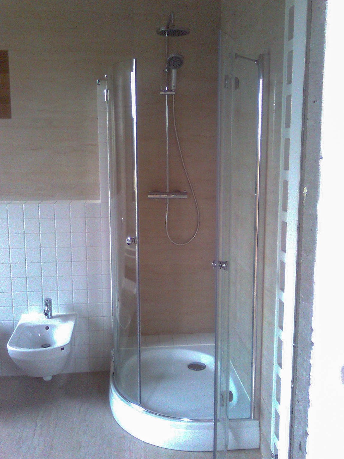 Kabina prysznicowa z zaokrąglonym frontem i szklanymi drzwiami, obok zamontowane białe bidet na tle kwadratowych płytek ściennych, widoczny fragment niedokończonej ściany.