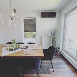 HVAC Business - Czarny klimatyzator ścienny zamontowany w nowoczesnym, jasnym wnętrzu jadalni, widoczny nad grzejnikiem i wysoką rośliną ozdobną, z drewnianym stołem nakrytym do posiłku.