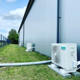 HVAC Business - Zewnętrzne jednostki klimatyzacji Hisense zamontowane na betonowych podstawach wzdłuż ściany hali przemysłowej, widoczne rury odprowadzające skropliny, tło to trawnik i ogrodzenie.