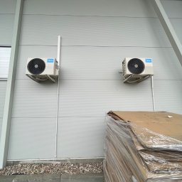 HVAC Business - Dwa klimatyzatory Midea zamontowane na metalowej ścianie hali przemysłowej, z widocznymi rurami odprowadzającymi, obok stosu kartonów owiniętych folią stretch.