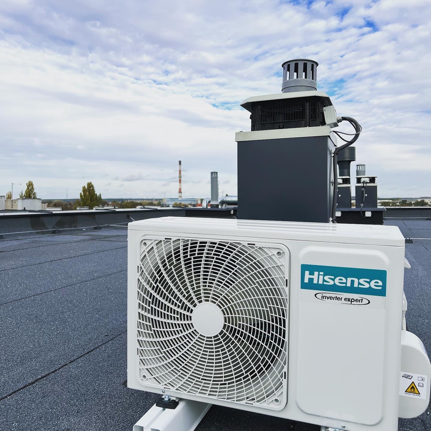 Biała jednostka zewnętrzna klimatyzacji Hisense Inverter Expert zamontowana na płaskim dachu z widocznymi elementami instalacji wentylacyjnej i kominem w tle, na tle pochmurnego nieba.