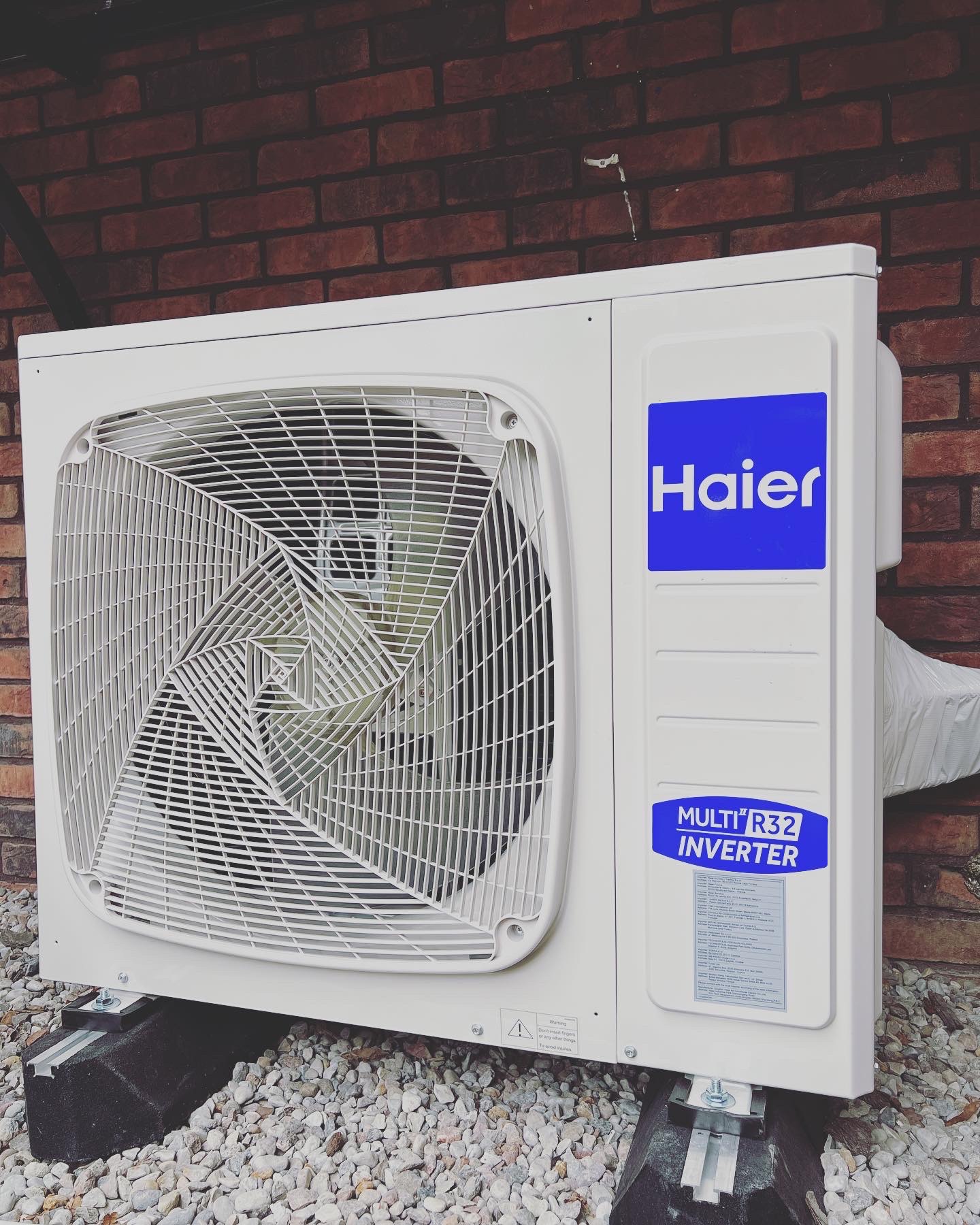 Zewnętrzna jednostka klimatyzacji Haier Multi R32 Inverter zamontowana na grafitowych wspornikach na tle ceglanej ściany i białego żwiru.