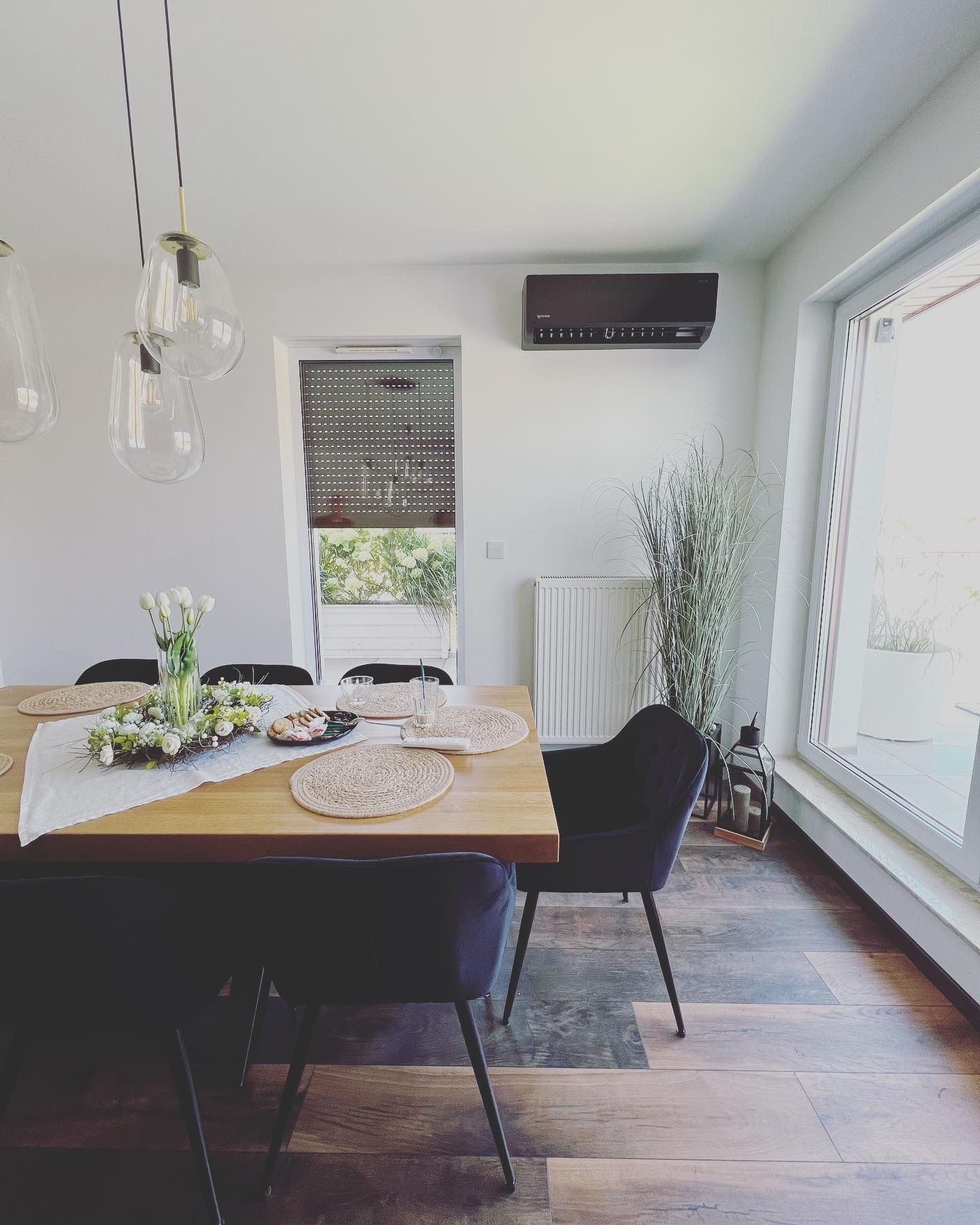 Czarny klimatyzator ścienny zamontowany w nowoczesnym, jasnym wnętrzu jadalni, widoczny nad grzejnikiem i wysoką rośliną ozdobną, z drewnianym stołem nakrytym do posiłku.