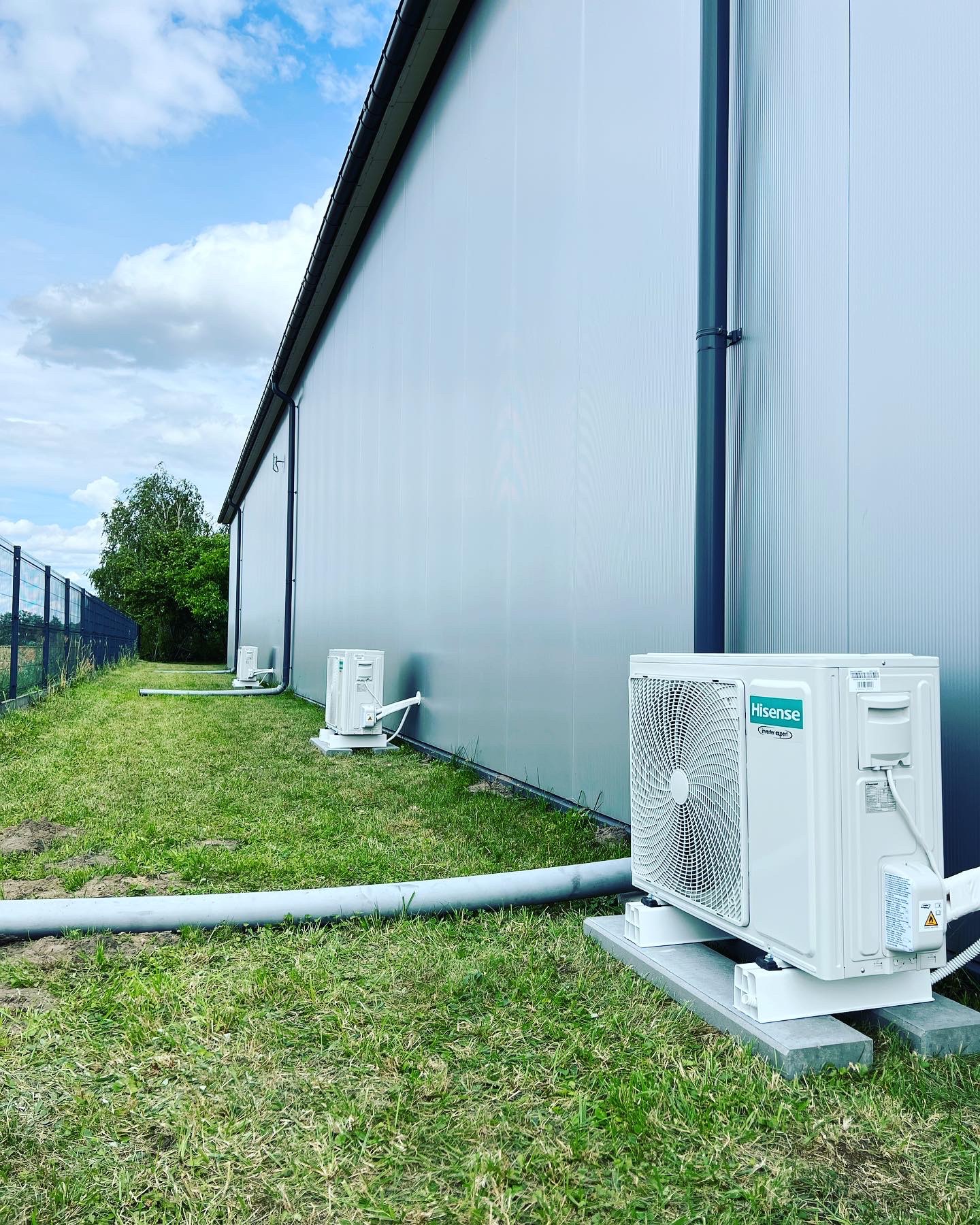 Zewnętrzne jednostki klimatyzacji Hisense zamontowane na betonowych podstawach wzdłuż ściany hali przemysłowej, widoczne rury odprowadzające skropliny, tło to trawnik i ogrodzenie.