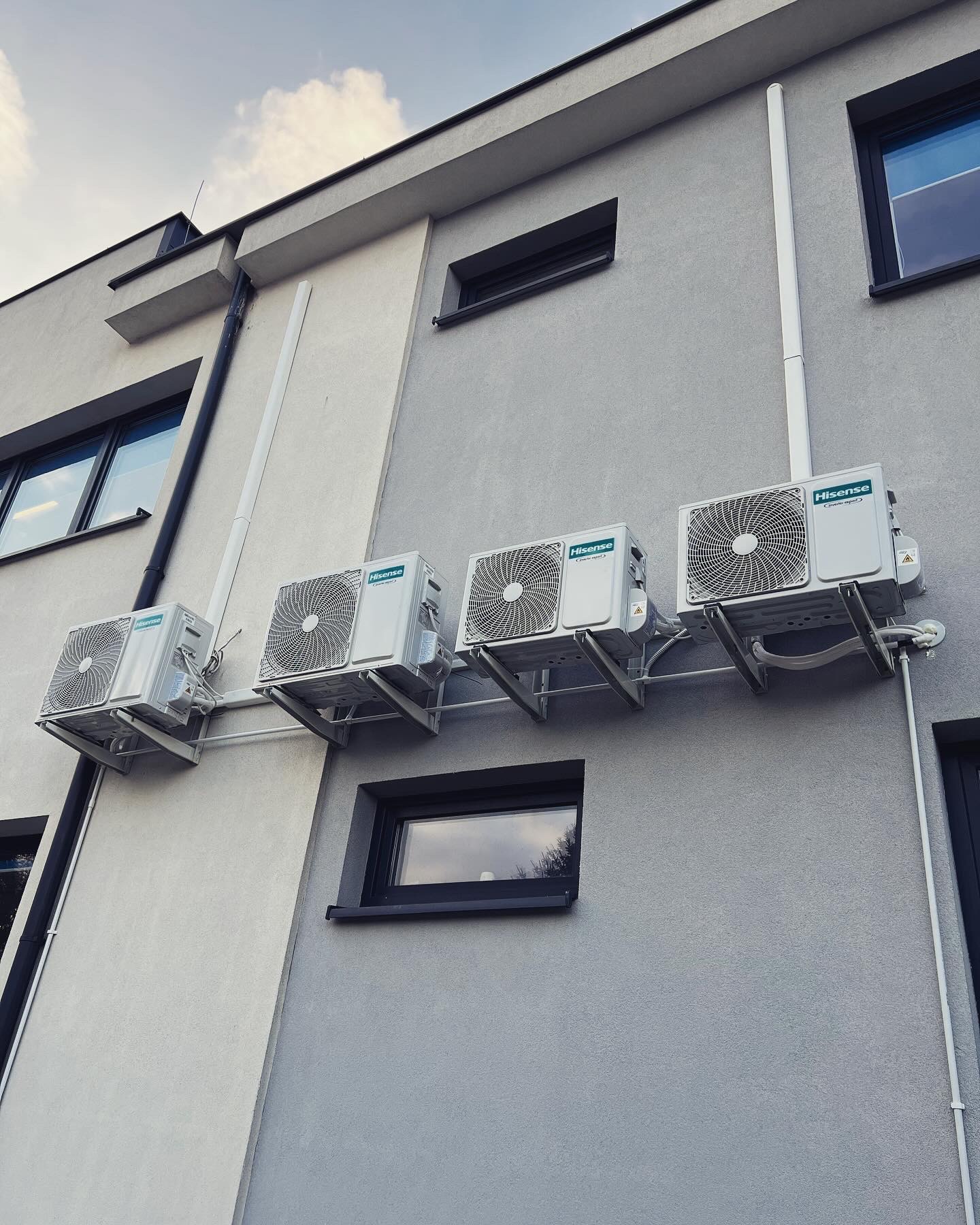 Zewnętrzne jednostki klimatyzacji marki Hisense zamontowane na elewacji budynku o szarej kolorystyce, widoczne rury odprowadzające skropliny i przewody elektryczne.
