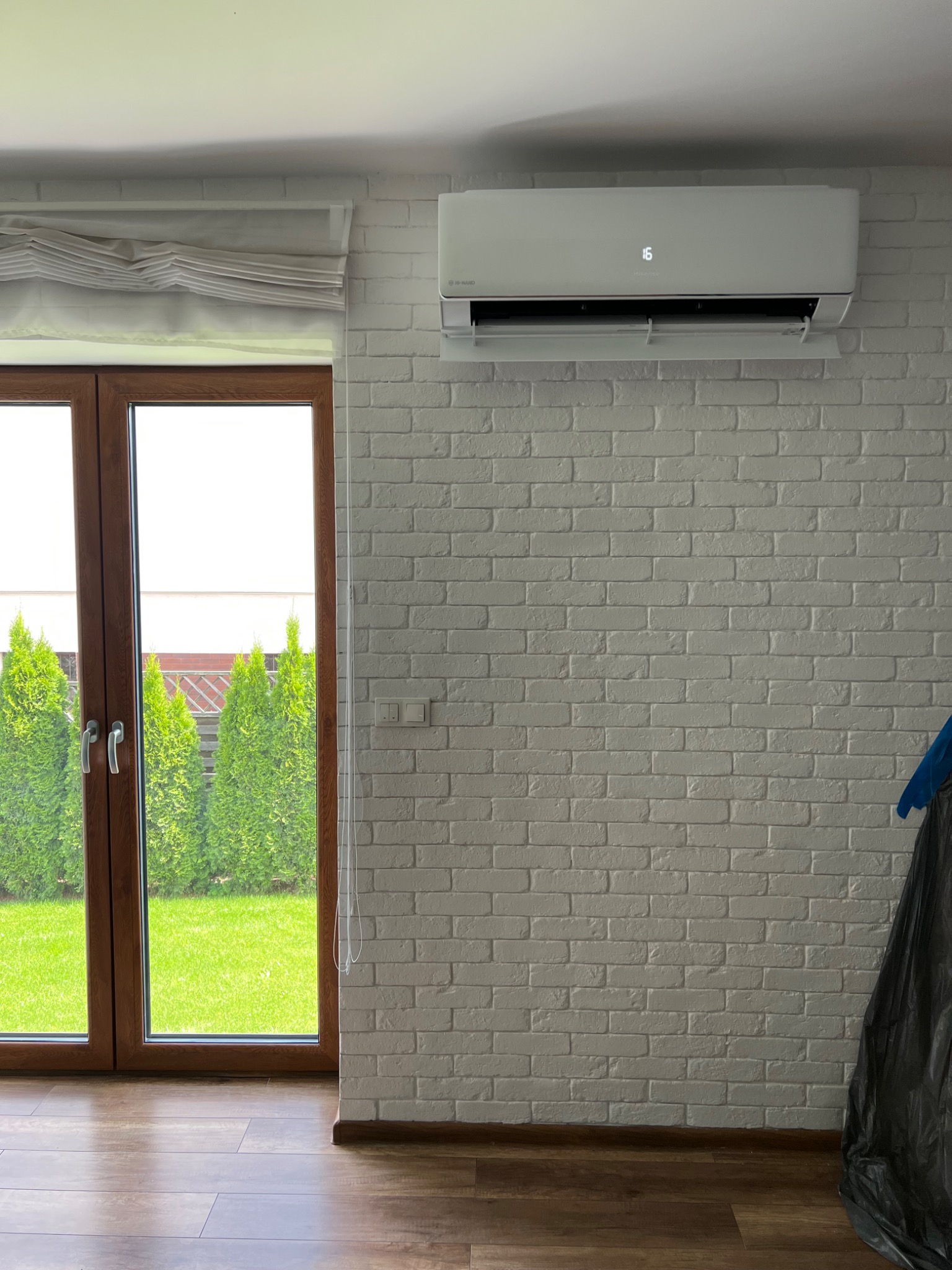 Biały klimatyzator ścienny na tle ściany z białej cegły, obok okna z widokiem na zielony ogród.