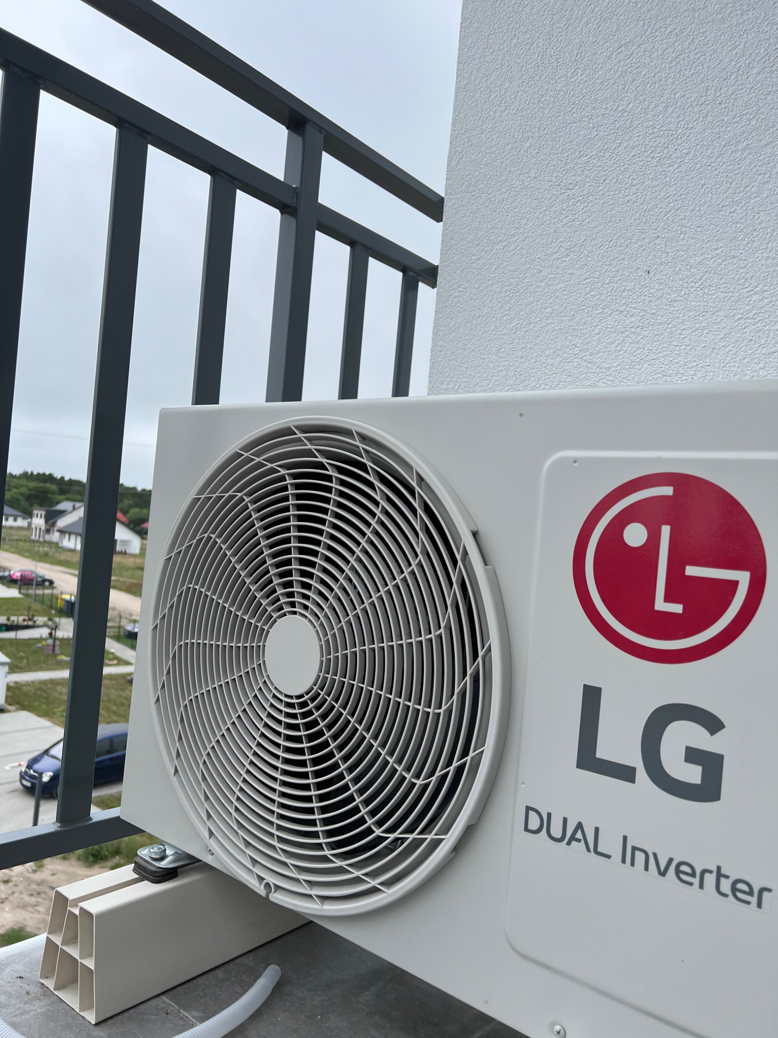 Zewnętrzna jednostka klimatyzacji LG Dual Inverter zamontowana na balkonie z widokiem na osiedle domów jednorodzinnych w pochmurny dzień.