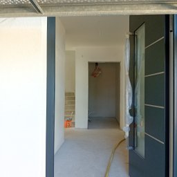 EG - Wnętrze w trakcie prac wykończeniowych: surowy beton na podłodze, pomalowane na biało ściany, częściowo osłonięte folią drzwi z czarną ramą i widoczne schody w tle. Pomarańczowe wiadro...