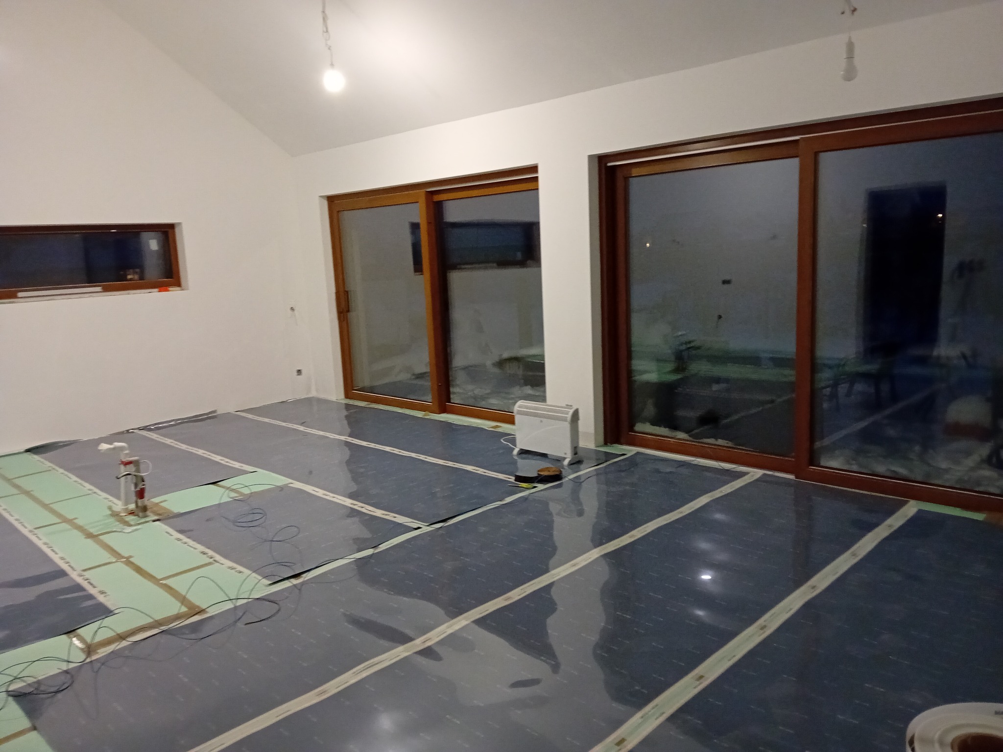 Przygotowanie podłoża pod ogrzewanie podłogowe: rozłożone maty grzewcze na zielonej izolacji, widoczne przewody, narzędzie pomiarowe i grzejnik elektryczny w pomieszczeniu z dużymi oknami.