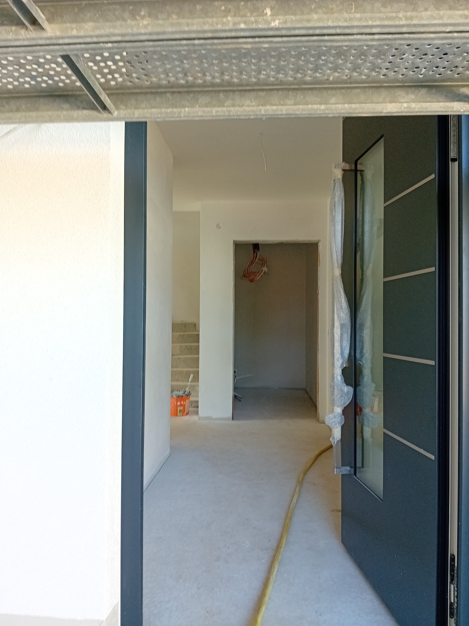 Wnętrze w trakcie prac wykończeniowych: surowy beton na podłodze, pomalowane na biało ściany, częściowo osłonięte folią drzwi z czarną ramą i widoczne schody w tle. Pomarańczowe wiadro...