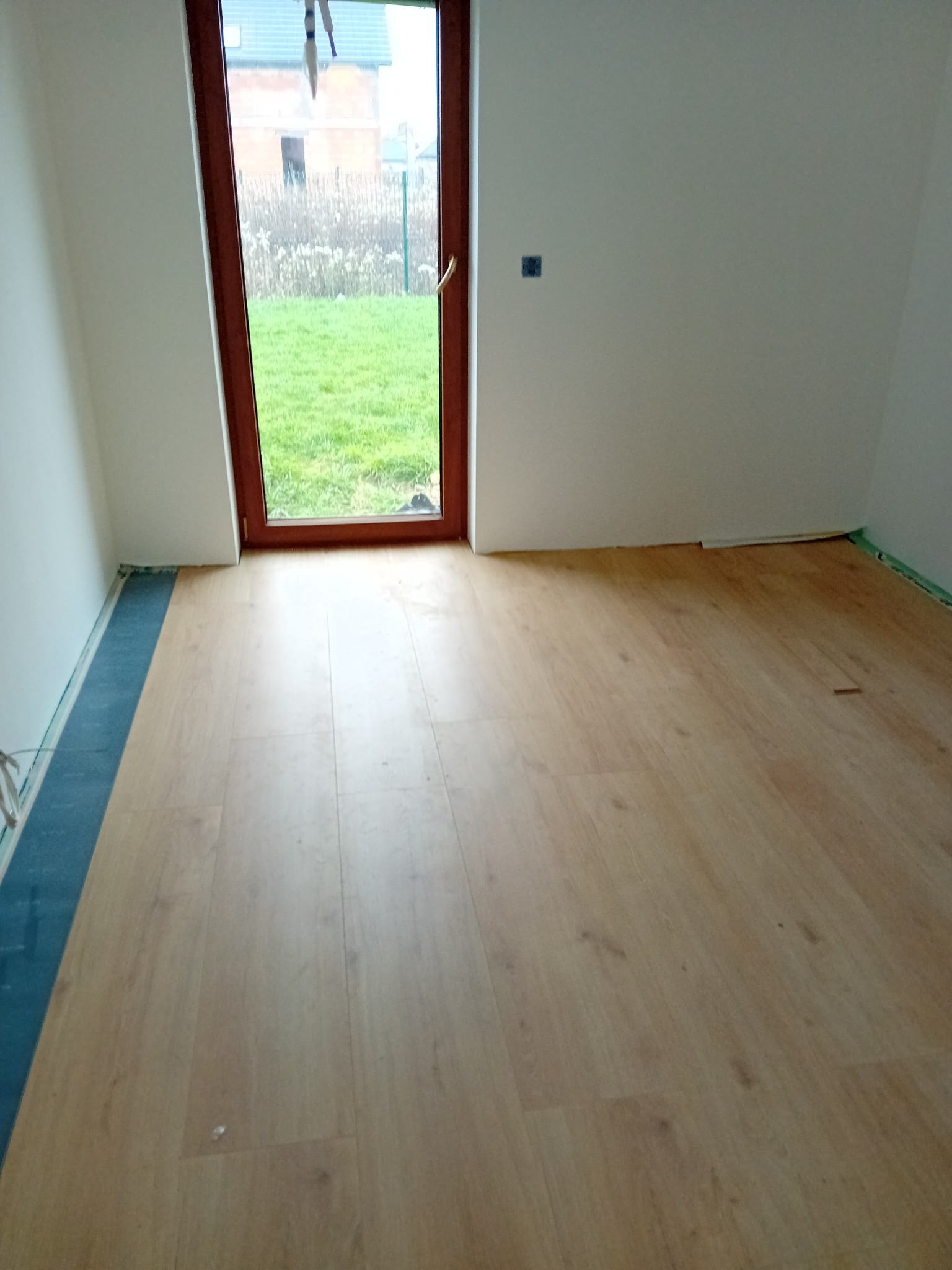 Jasne panele podłogowe ułożone w pokoju z białymi ścianami, widoczne okno z brązową ramą i zielony trawnik na zewnątrz, listwy przypodłogowe zabezpieczone taśmą malarską.