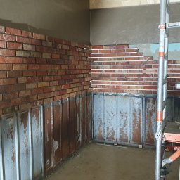 NewHome - Wnętrze w trakcie wykańczania z częściowo położoną cegłą dekoracyjną na ścianach, widoczne listwy dystansowe, rusztowanie i metalowe panele ścienne na dole.