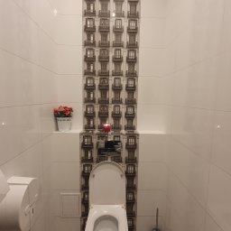 NewHome - Wykończona toaleta z białymi płytkami, centralny pas dekoracyjny z motywem okien kamienicy, drewnopodobna podłoga, wisząca muszla klozetowa i czerwona świeczka na półce.