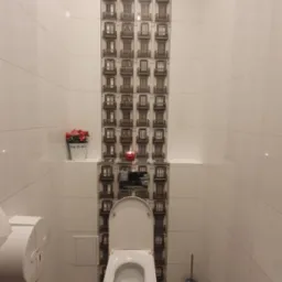 Wykończona toaleta z białymi płytkami, centralny pas dekoracyjny z motywem okien kamienicy, drewnopodobna podłoga, wisząca muszla klozetowa i czerwona świeczka na półce.