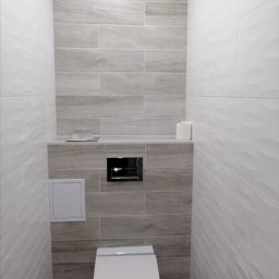 NewHome - Wąska toaleta wykończona szarymi płytkami imitującymi drewno na ścianie za muszlą klozetową, kontrastującymi z białymi płytkami o falistej strukturze po bokach, z czarnym przyciskiem spłukującym...