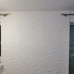 NewHome - Ściana wykończona białymi panelami dekoracyjnymi imitującymi kamień, z widocznymi karniszami po bokach, przy dolnej krawędzi widoczna butelka PET.