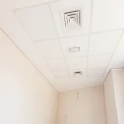 Wnętrze pokoju w trakcie wykańczania, widoczny sufit podwieszany kasetonowy z kratkami wentylacyjnymi i czujnikiem dymu, ściany przygotowane pod malowanie z widocznymi śladami po instalacjach.