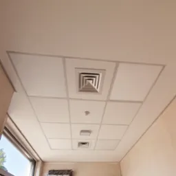 Kasetonowy sufit podwieszany z kratką wentylacyjną i czujnikiem dymu, widoczny klimatyzator w folii ochronnej i drabina w rogu pomieszczenia.