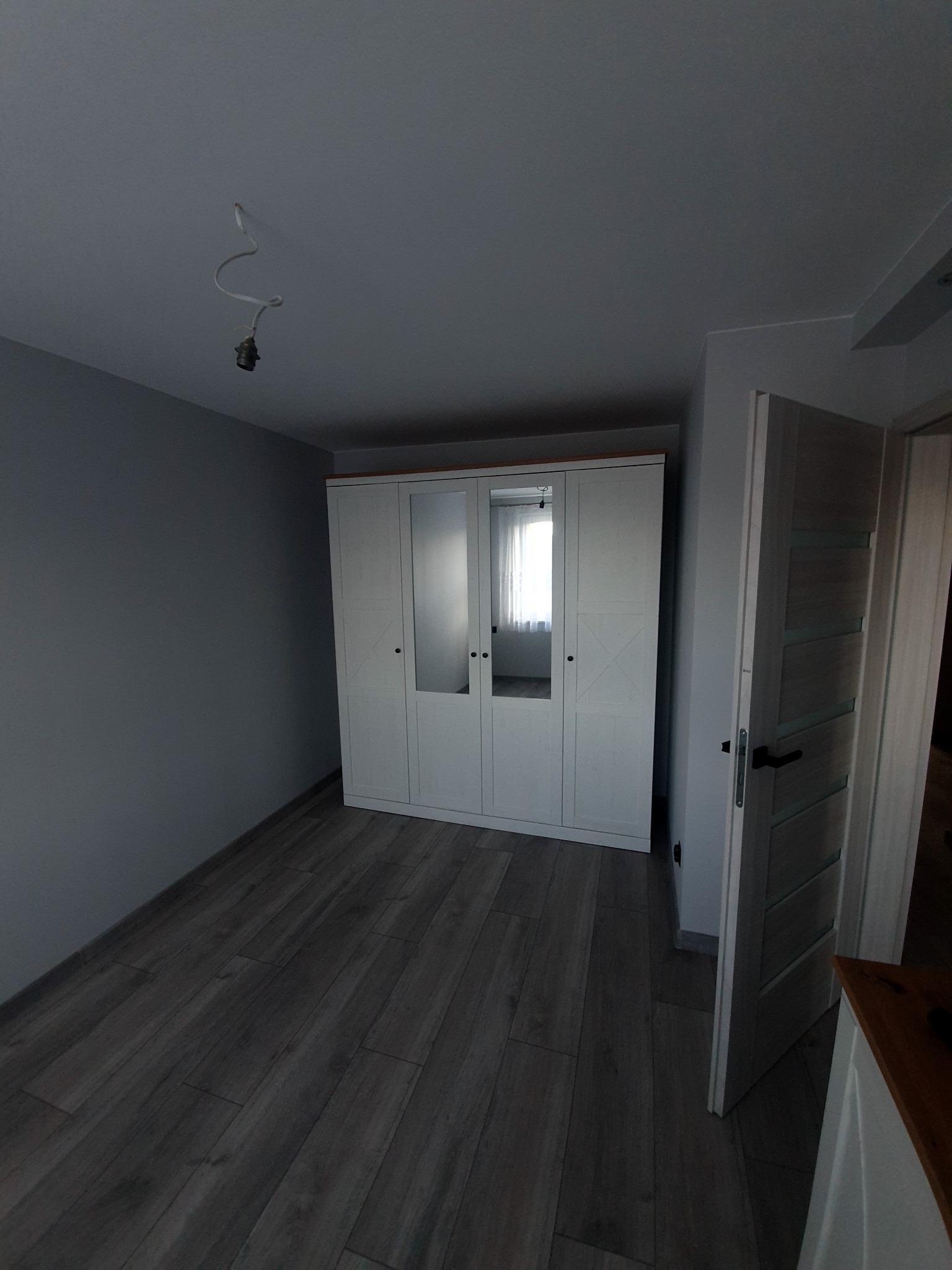 Pusty pokój z szarą ścianą, panelami podłogowymi w odcieniach brązu i białym, trzydrzwiowym kredensem z lustrami na środkowych drzwiach. Na suficie widoczna wisząca oprawka bez żarówki.