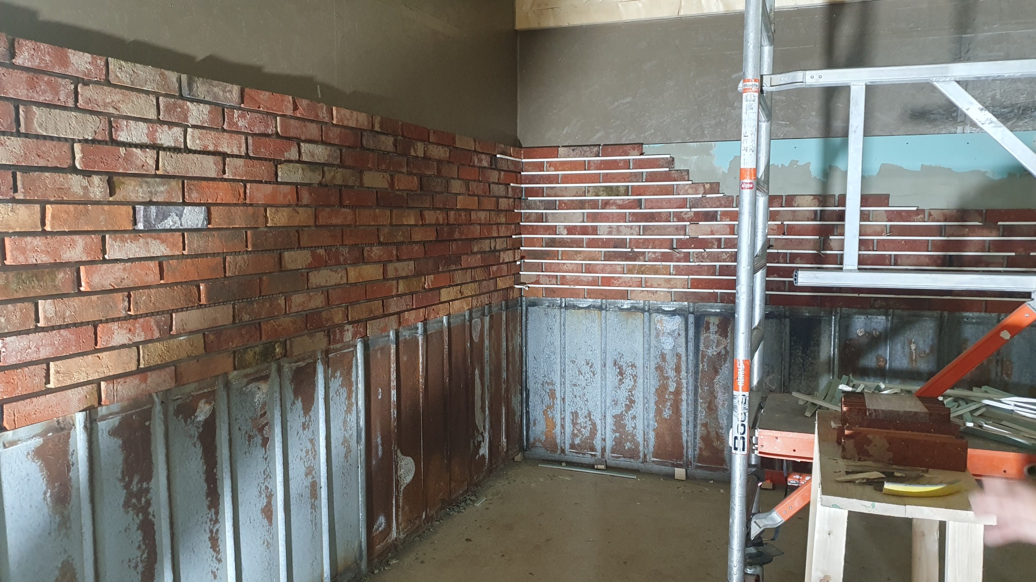 Wnętrze w trakcie wykańczania z częściowo położoną cegłą dekoracyjną na ścianach, widoczne listwy dystansowe, rusztowanie i metalowe panele ścienne na dole.