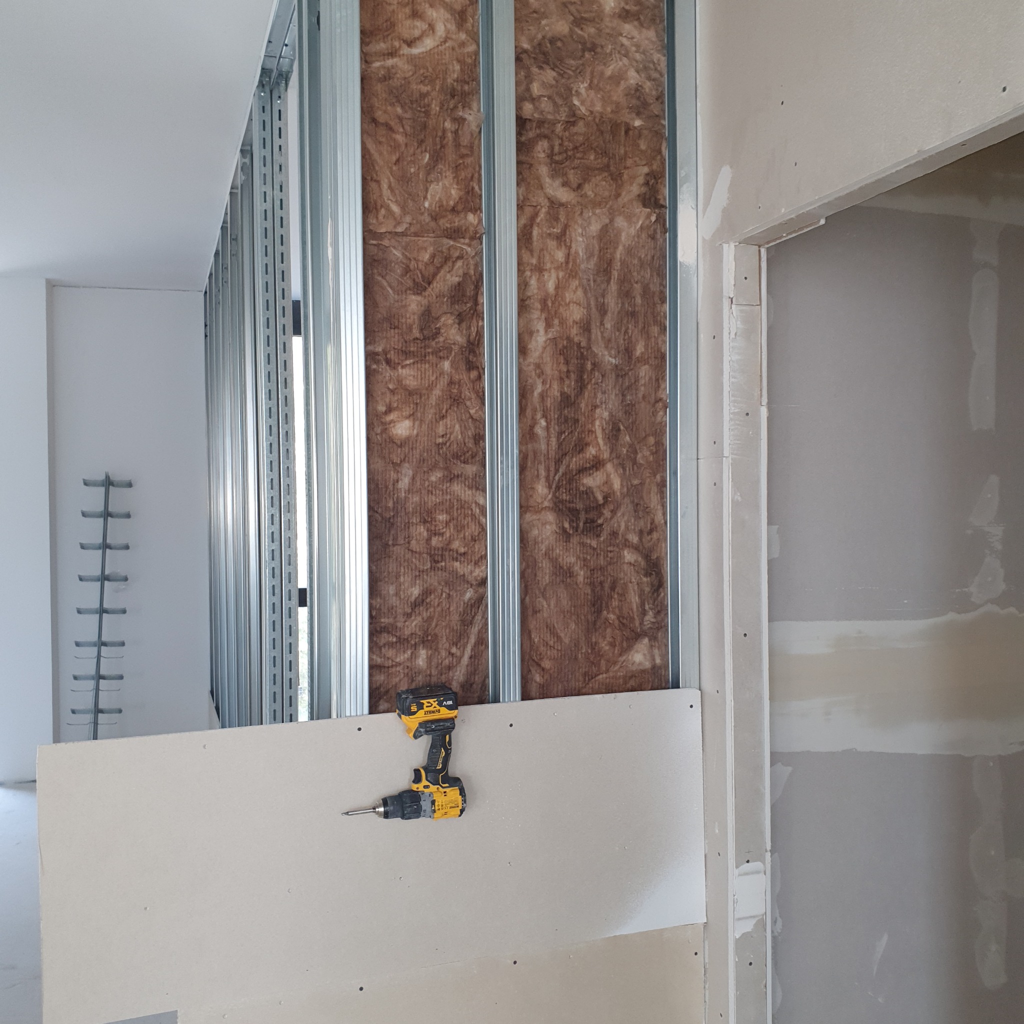 Montaż płyty gipsowo-kartonowej na metalowym stelażu z izolacją z wełny mineralnej, wiertarka akumulatorowa DeWalt leży na płycie.