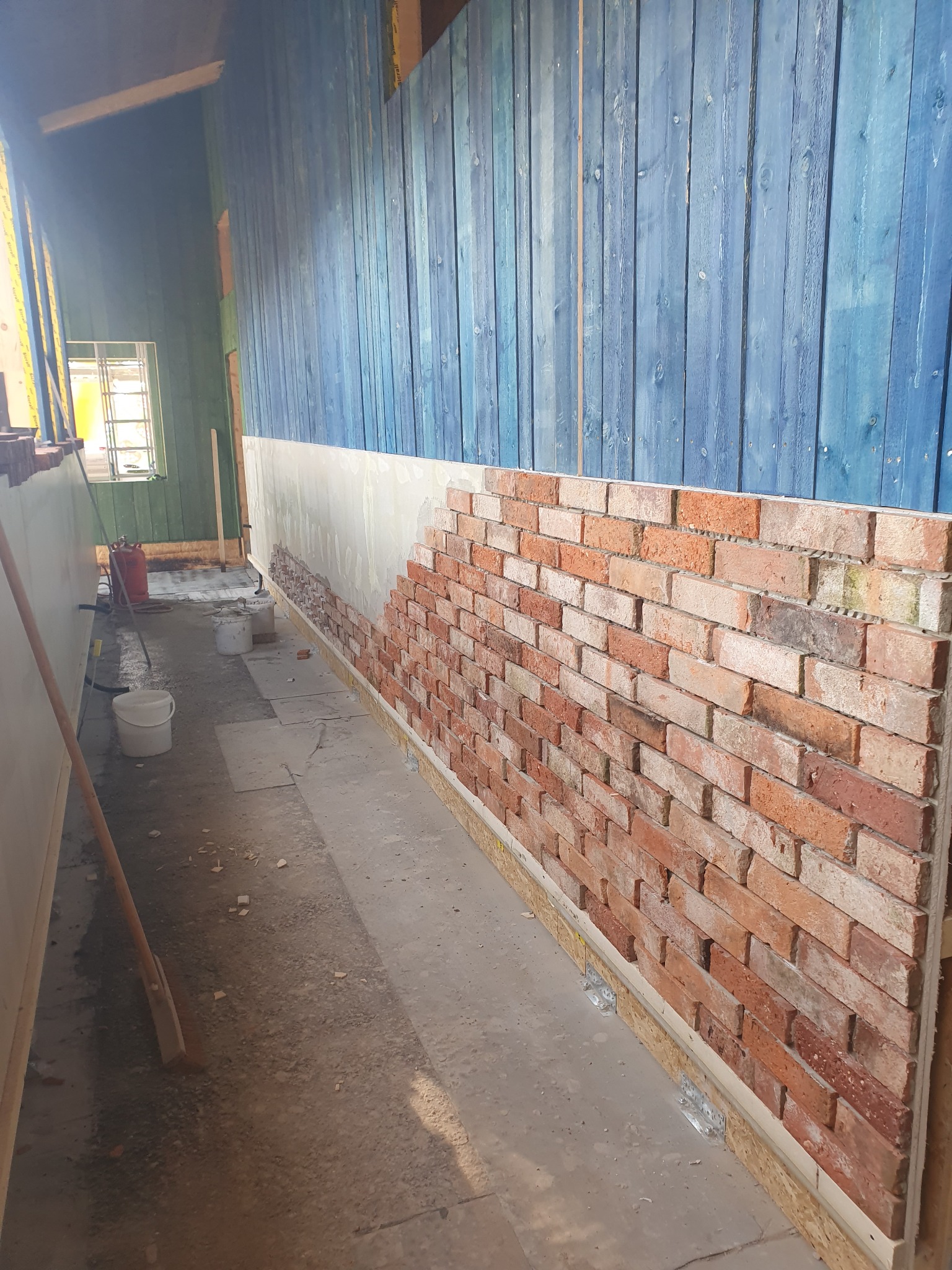 Montaż dekoracyjnej ściany z cegieł na płycie OSB wewnątrz pomieszczenia z niebieskimi, drewnianymi panelami ściennymi. Widoczne elementy mocujące i etap prac w trakcie realizacji.