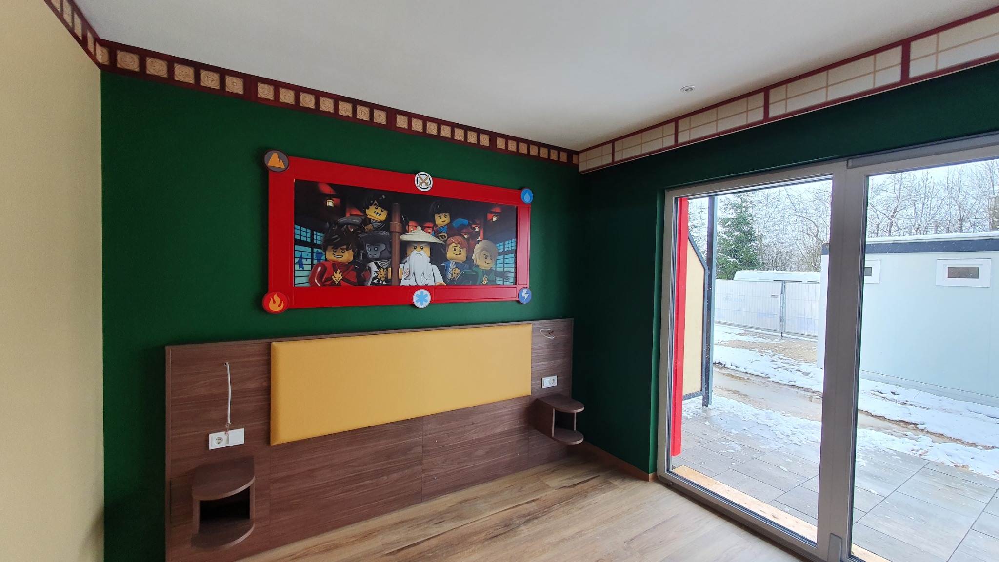 Pokój tematyczny z zielonymi ścianami, dekoracyjną bordiurą, łóżkiem z drewnianym wezgłowiem i żółtym panelem oraz dużym obrazem Lego Ninjago w czerwonej ramie.