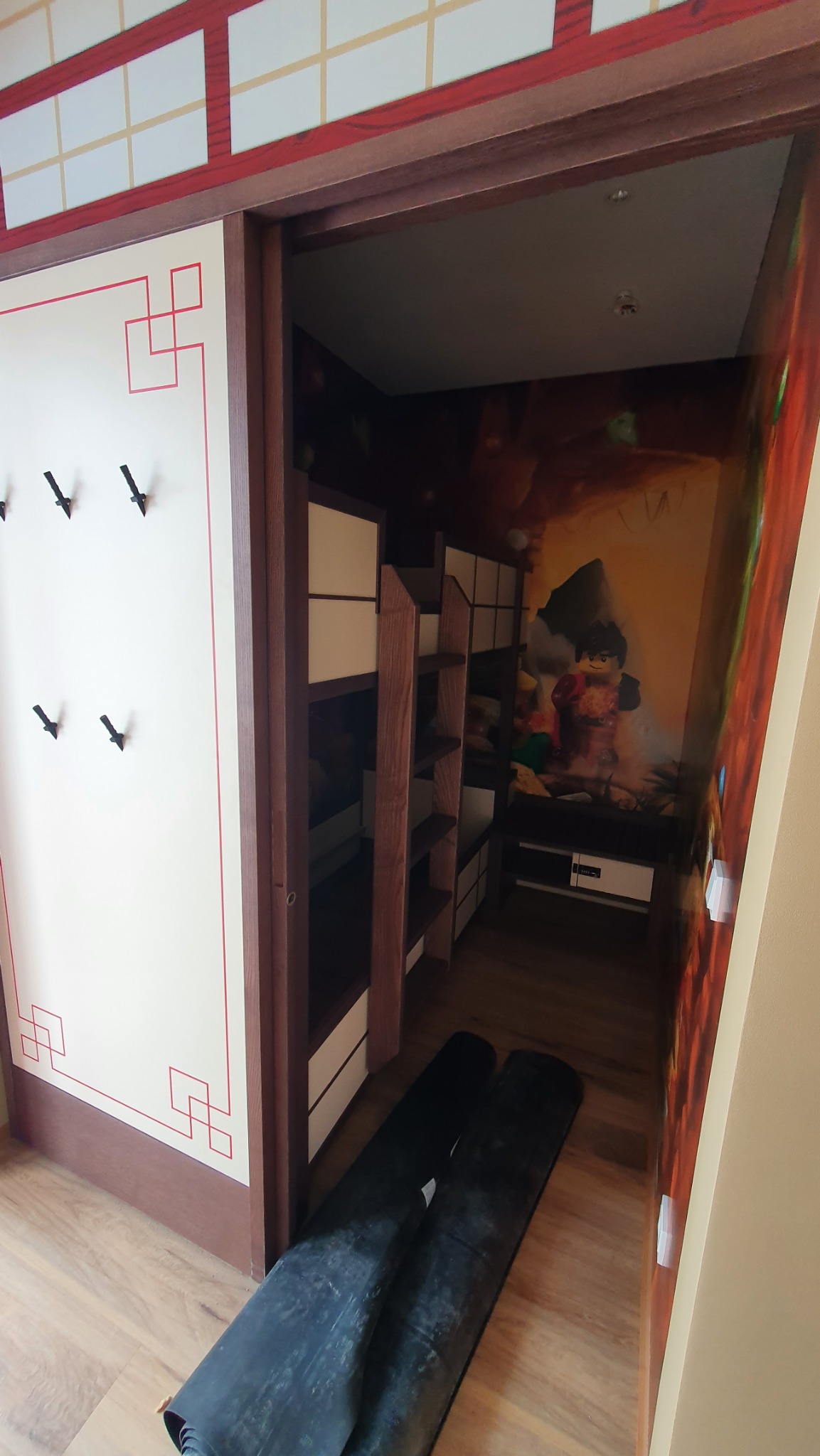 Pokój dziecięcy z motywem Lego Ninjago, ściana z wieszakami w stylu japońskim, widoczne dwie rolki czarnej folii na podłodze.