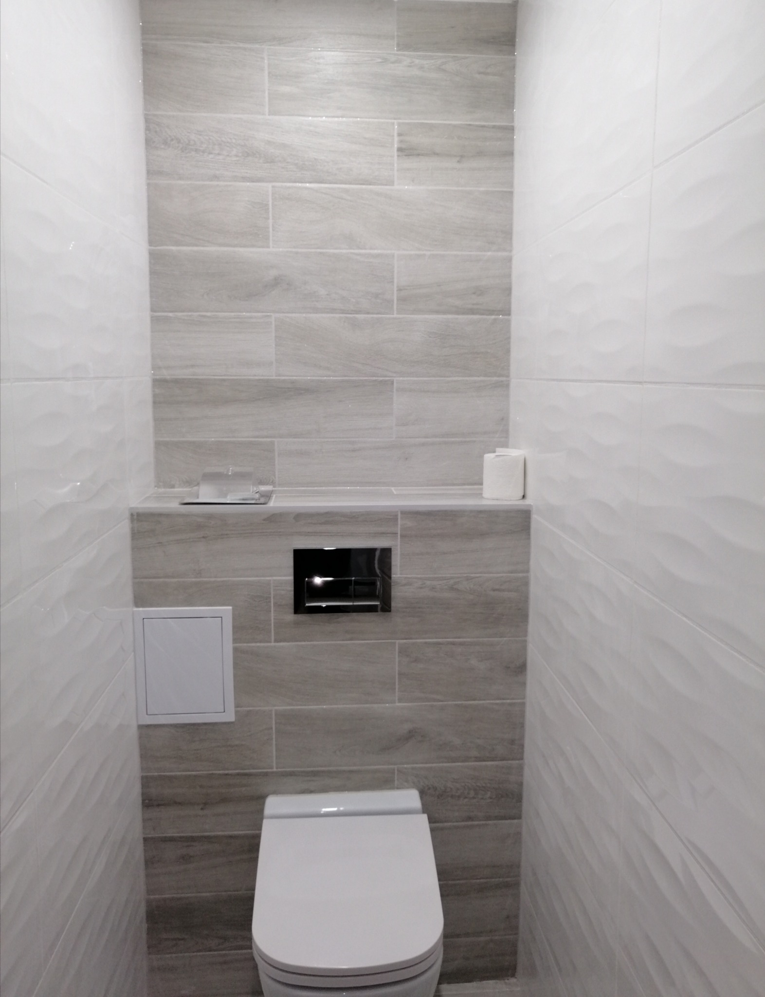 Wąska toaleta wykończona szarymi płytkami imitującymi drewno na ścianie za muszlą klozetową, kontrastującymi z białymi płytkami o falistej strukturze po bokach, z czarnym przyciskiem spłukującym...