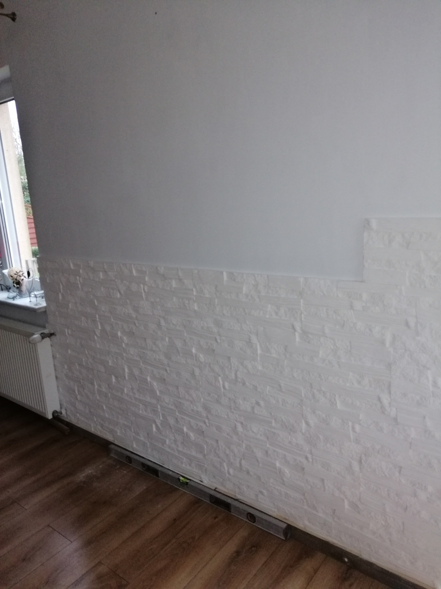 Fragment ściany z częściowo położoną dekoracyjną, białą okładziną imitującą kamień, widoczny poziomica na podłodze, górna część ściany pomalowana na biało.