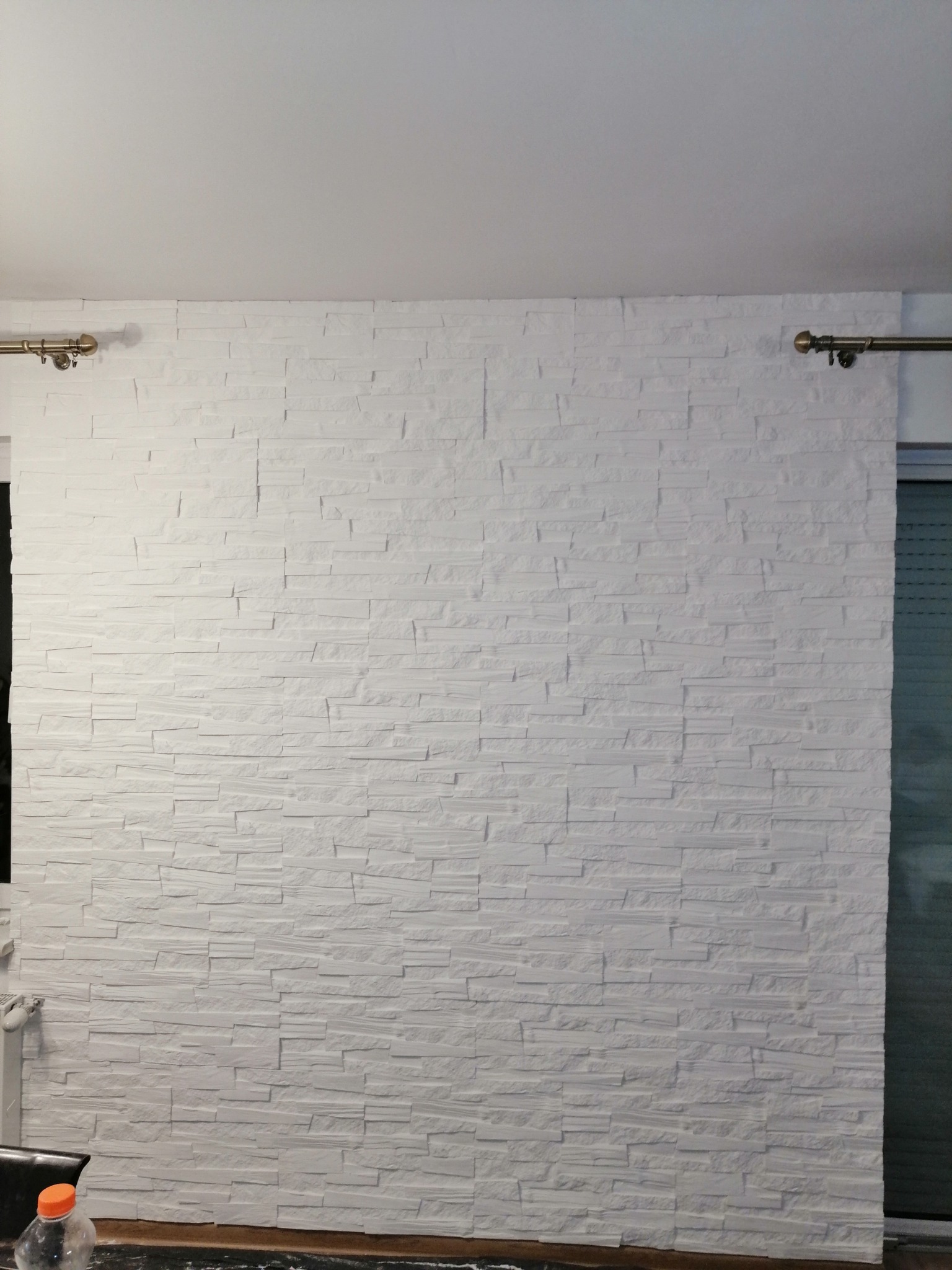 Ściana wykończona białymi panelami dekoracyjnymi imitującymi kamień, z widocznymi karniszami po bokach, przy dolnej krawędzi widoczna butelka PET.