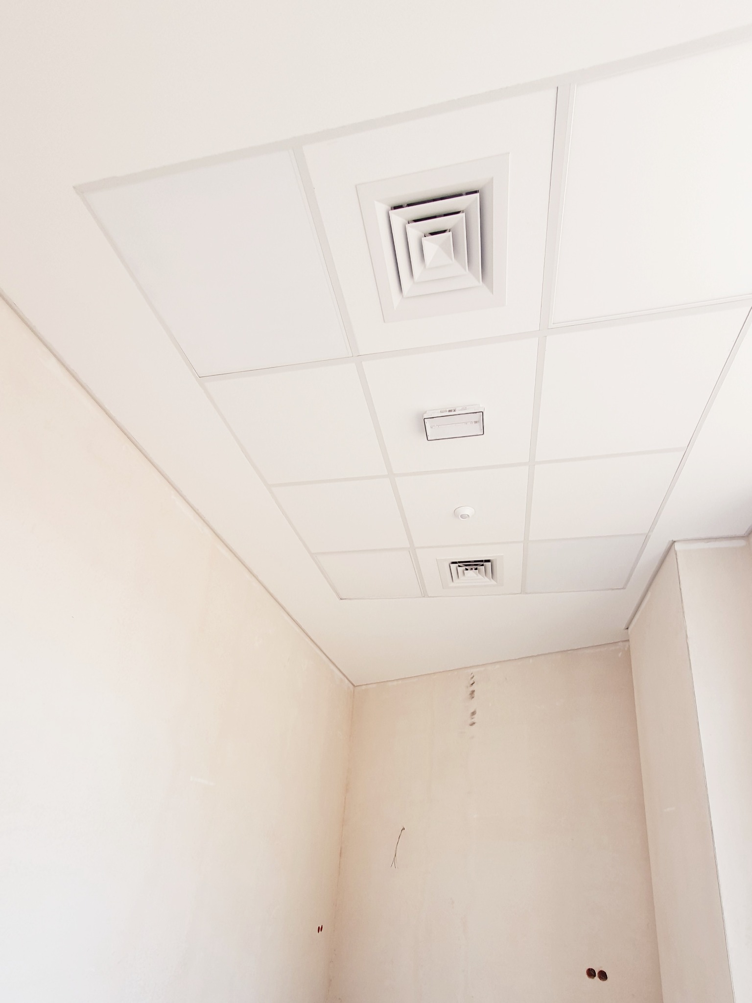 Wnętrze pokoju w trakcie wykańczania, widoczny sufit podwieszany kasetonowy z kratkami wentylacyjnymi i czujnikiem dymu, ściany przygotowane pod malowanie z widocznymi śladami po instalacjach.