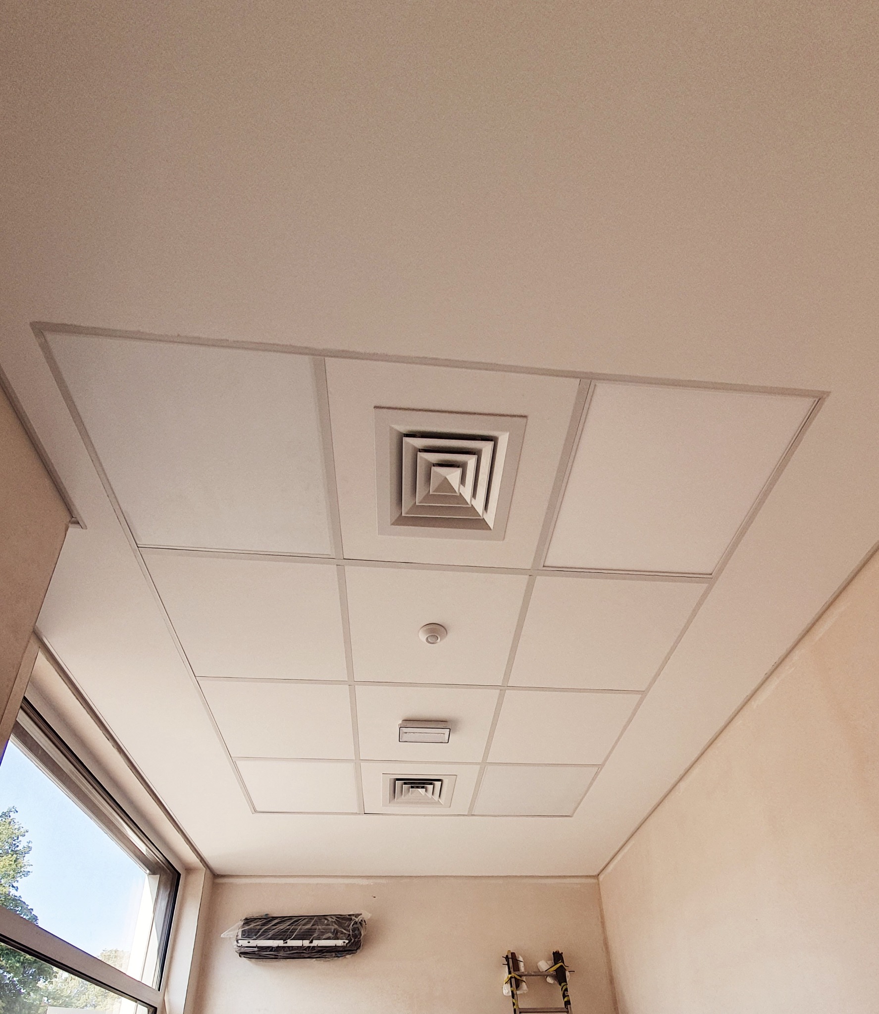 Kasetonowy sufit podwieszany z kratką wentylacyjną i czujnikiem dymu, widoczny klimatyzator w folii ochronnej i drabina w rogu pomieszczenia.